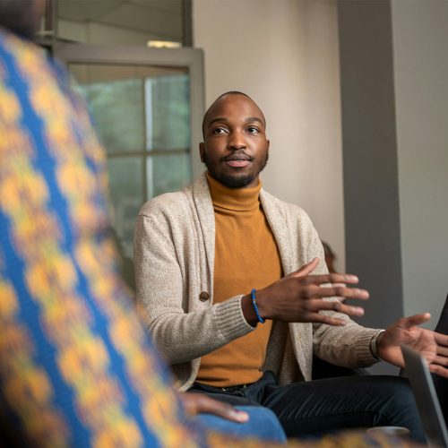 A man in a mustard sweater and beige cardigan is speaking and gesturing with his hands in an office setting, sitting with others in discussion.
