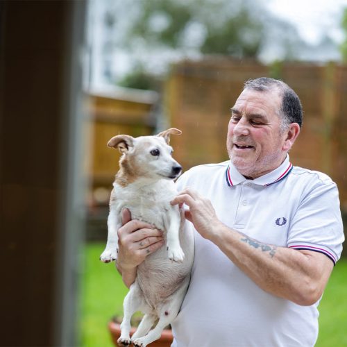 A man holds a small dog, smiling in a garden setting with blurred foliage and fencing in the background.