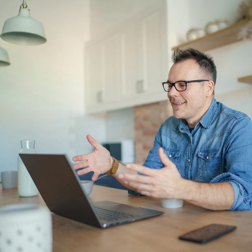 A man in glasses and a denim shirt gestures while talking in front of an open laptop in a modern kitchen with light fixtures and shelves in the background.