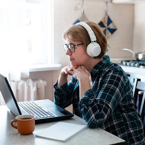 A person wearing headphones and a plaid shirt sits at a table, focused on a laptop. A notebook and mug are nearby in a bright kitchen.
