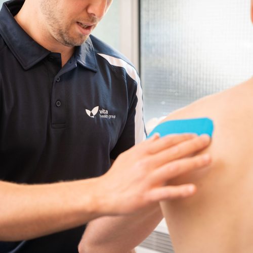 A healthcare professional applies a blue therapeutic tape to a patient's back shoulder. The professional wears a Vita Health Group polo shirt, indicating a clinical setting.