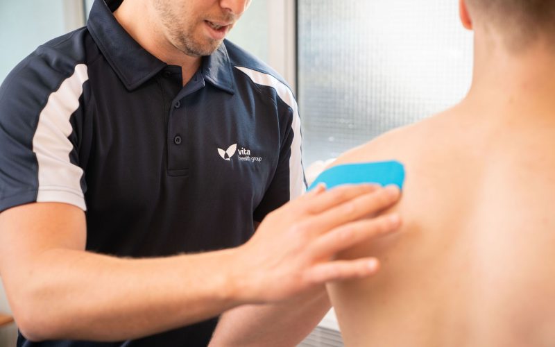 A healthcare professional applies a blue therapeutic tape to a patient's back shoulder. The professional wears a Vita Health Group polo shirt, indicating a clinical setting.