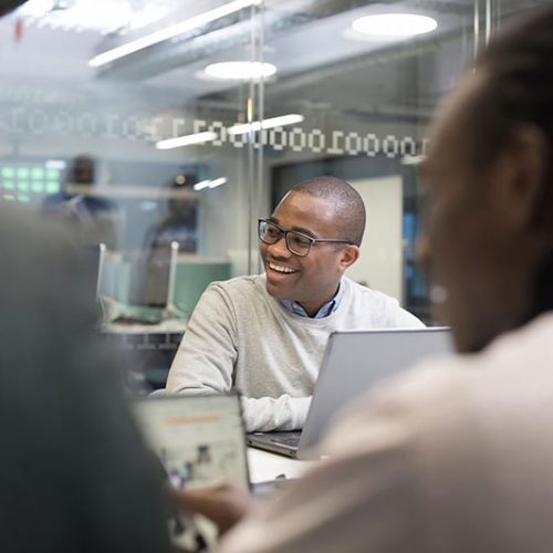 A person sits at a desk, smiling and discussing with colleagues in a modern, glass-walled office setting, with a laptop open in front of them.