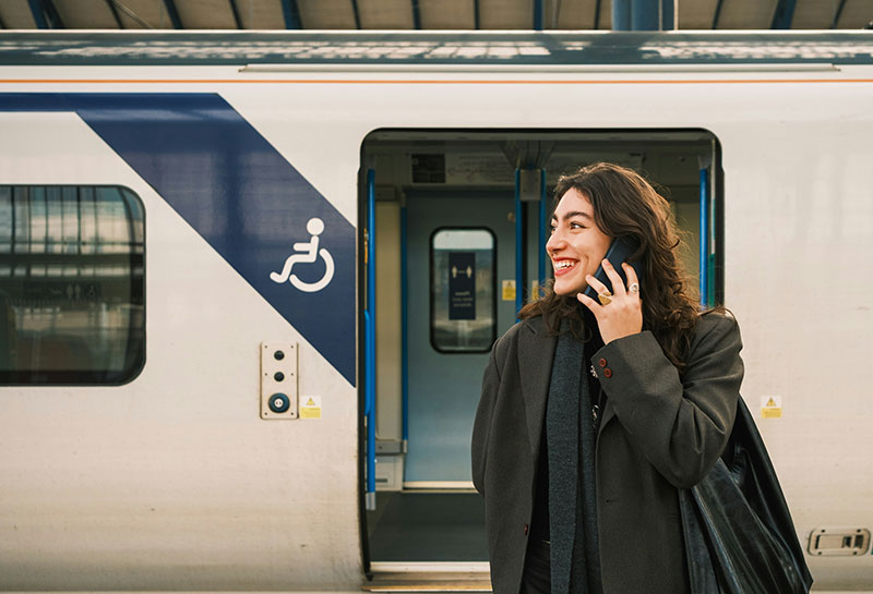 A person stands on a train platform, wearing a black coat and looking to the side. Behind them, a train with a partially visible logo is stationed.