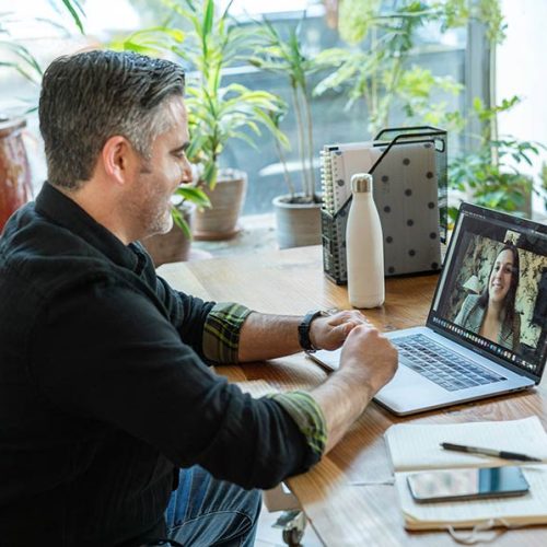 A man sits at a wooden desk engaging in a video call on a laptop. The room has large windows with plants visible in the background, creating a bright, natural atmosphere.
