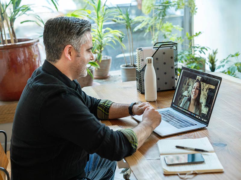 A man sits at a wooden desk engaging in a video call on a laptop. The room has large windows with plants visible in the background, creating a bright, natural atmosphere.