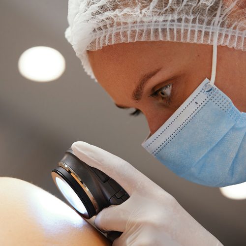 A healthcare professional, wearing a mask and hairnet, examines a patient's skin with a dermatoscope in a clinical setting, indicating a focus on skin health and medical assessment.