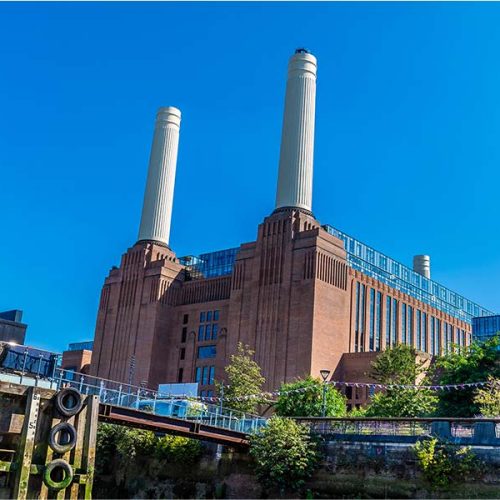 A historic industrial building with two tall chimneys stands against a clear blue sky, surrounded by greenery and adjacent modern structures. A bridge crosses a waterway in the foreground.