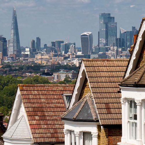 Rooftops of suburban houses with brown tiles in the foreground, set against a backdrop of a city skyline featuring modern skyscrapers under a partly cloudy sky.