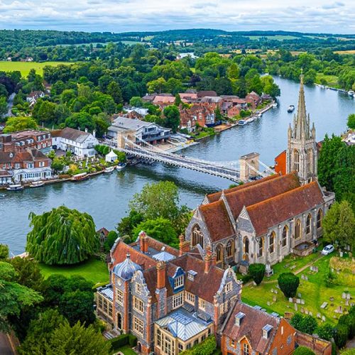 Aerial view of a historic church with a tall spire next to a river, lush greenery, and a bridge connecting charming houses in a picturesque countryside setting.