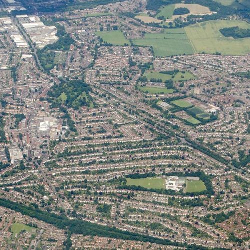 Aerial view of a densely populated area, featuring rows of houses interspersed with green spaces and fields, surrounded by trees and larger patches of greenery in the background.