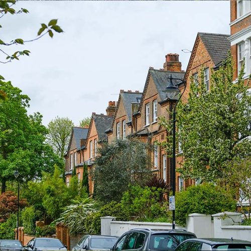 A row of traditional brick houses lines a residential street, surrounded by lush green trees and shrubs. Parked cars are visible along the street under a cloudy sky.