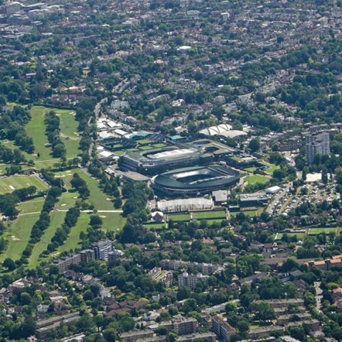 Aerial view of a city landscape featuring sports complexes with open fields and tennis courts. Surrounded by residential buildings and green spaces, the scene captures a lively urban environment.