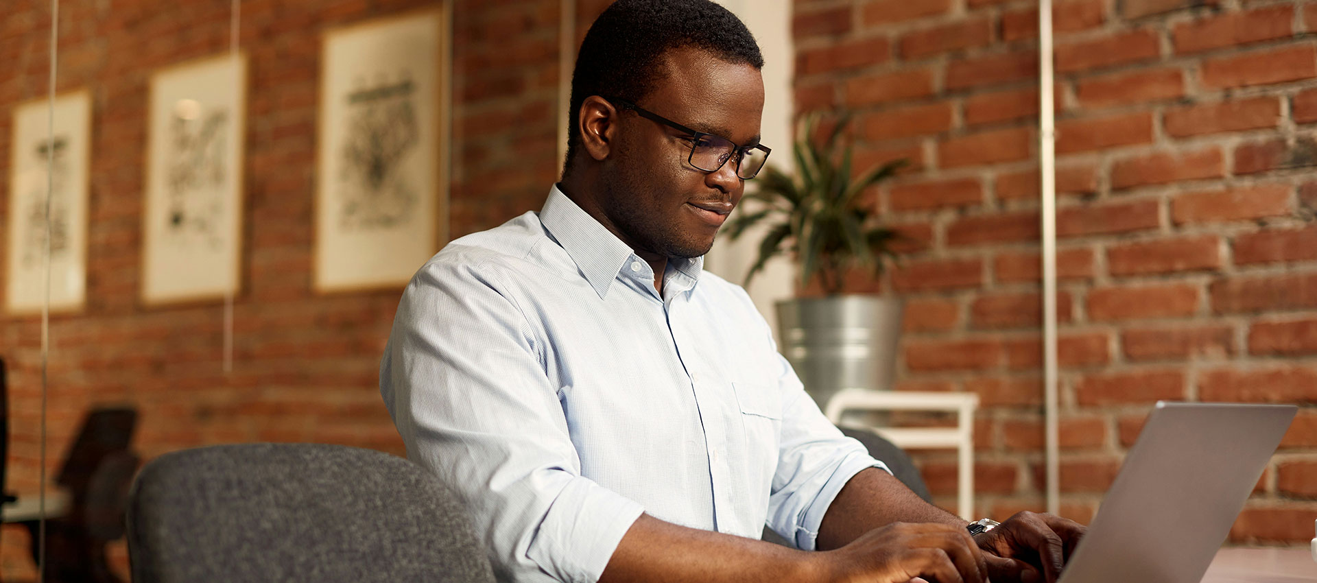 A person in a light blue shirt is typing on a laptop in an office with a brick wall and framed art in the background.