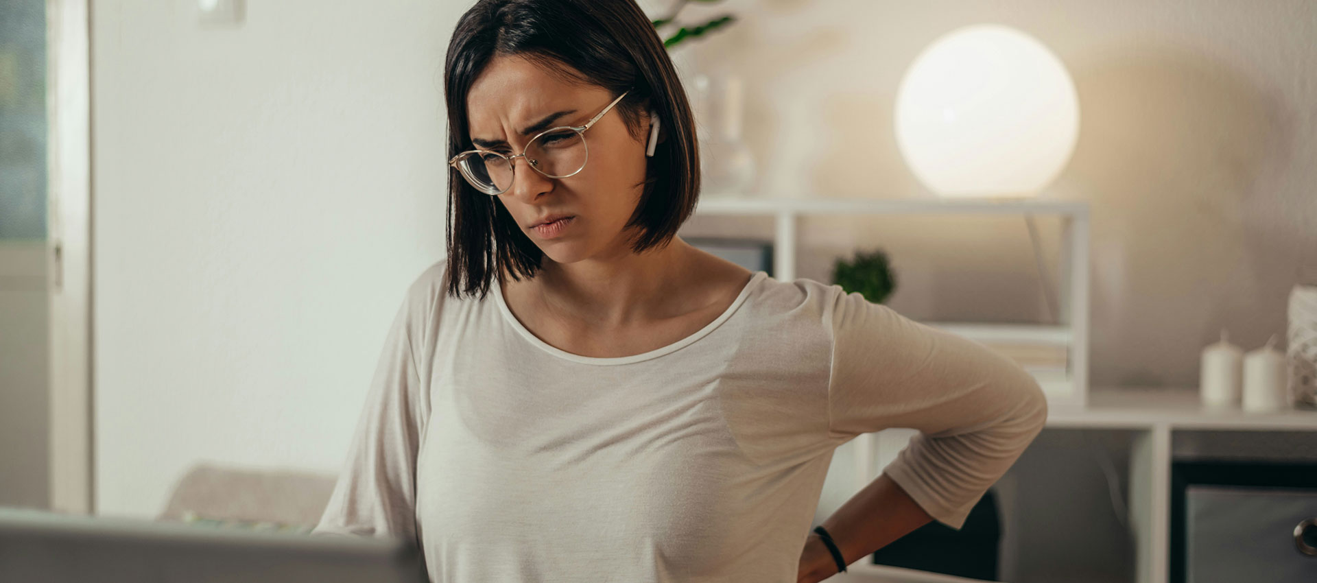 A woman wearing glasses sits at a desk, holding her lower back in discomfort, in a softly lit room with a glowing lamp and shelving in the background.