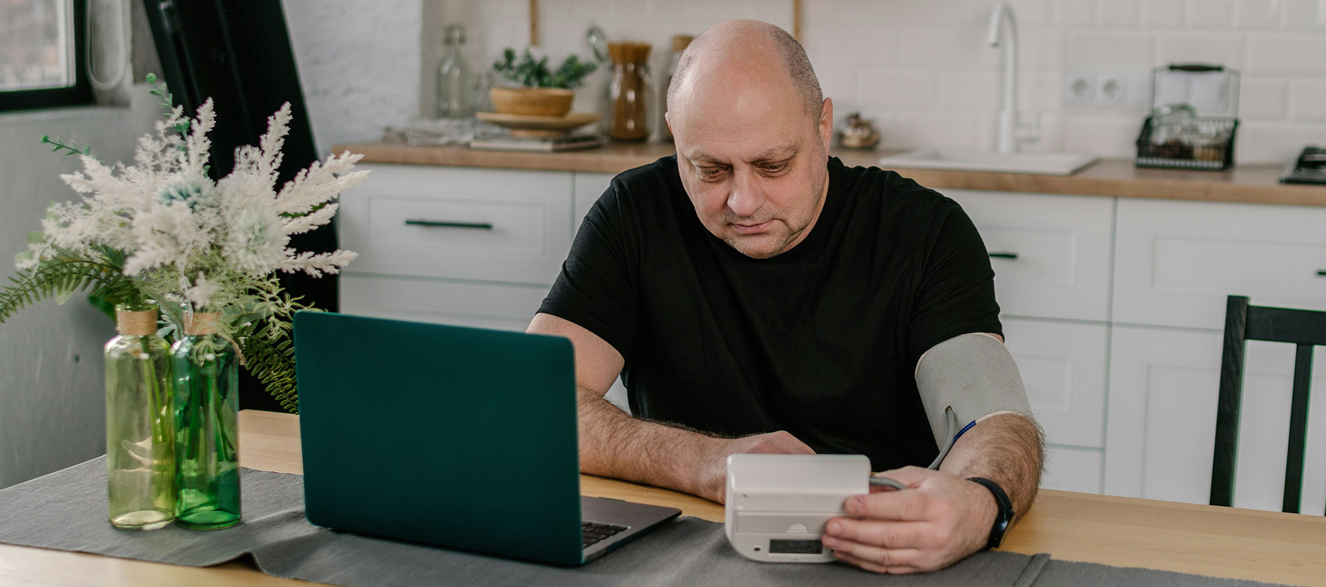 A man with a cardiovascular disease checks his blood pressure using a monitor at a kitchen table, with a laptop nearby and decorative plants in the background.