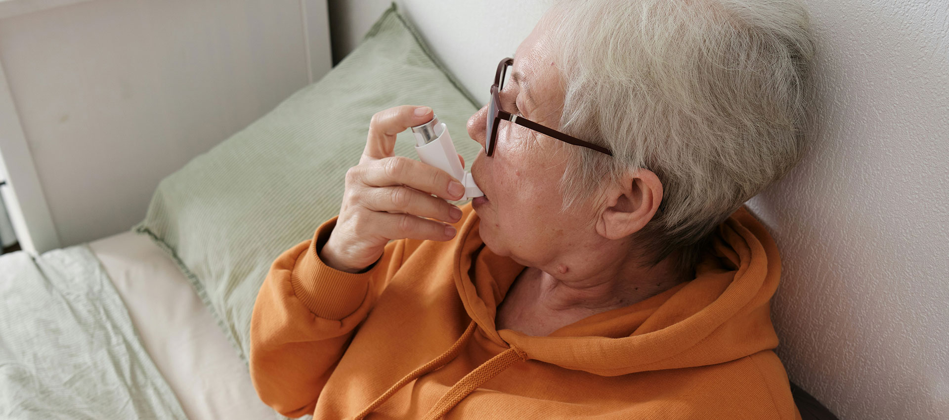 An older adult wearing glasses uses an inhaler while sitting against a white wall, dressed in an orange hoodie, with cushions nearby.
