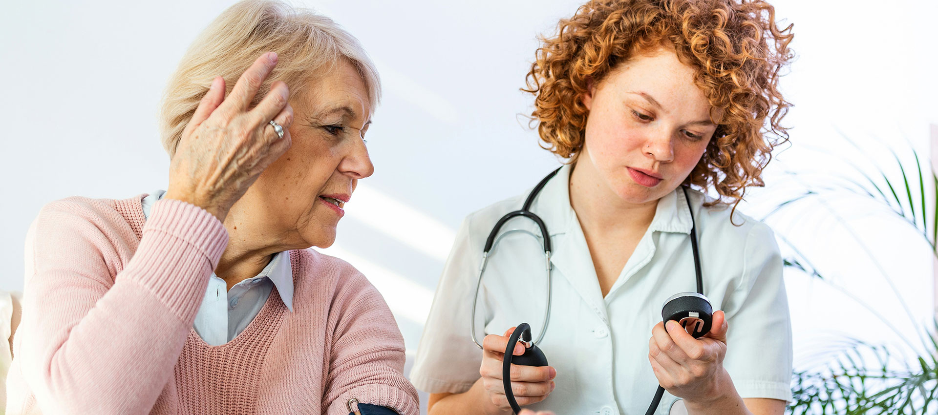 A healthcare professional examines a blood pressure monitor while a patient with a long term condition, seated beside her, listens attentively. They are indoors, with a potted plant in the background.