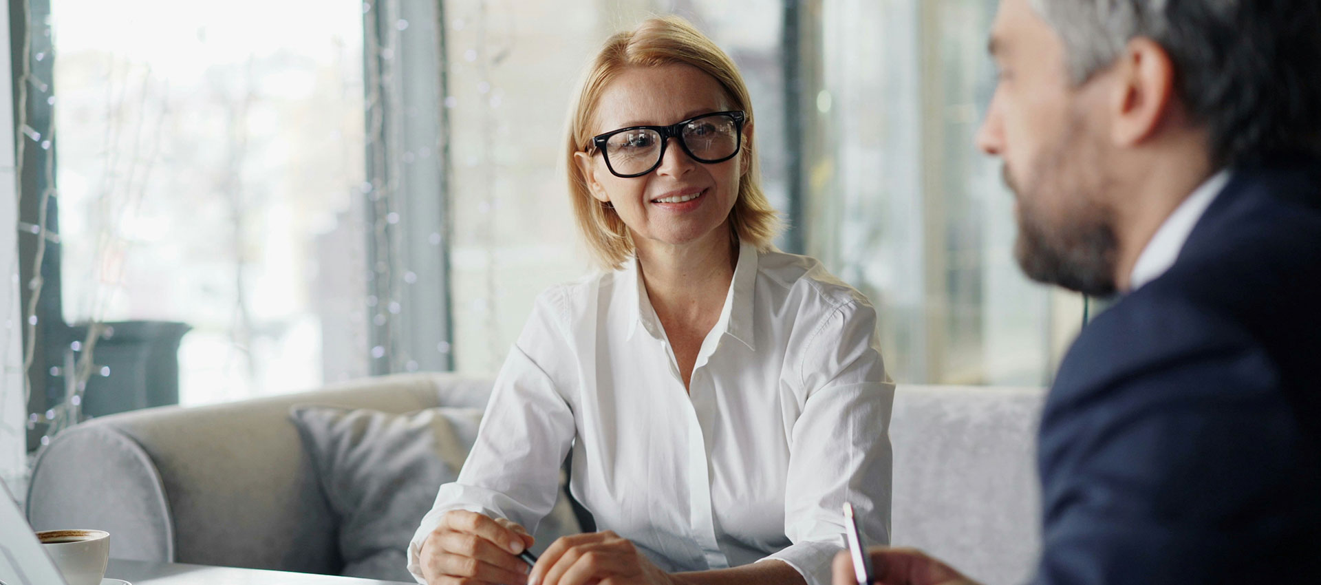 A woman wearing glasses smiles while sitting on a sofa, engaged in conversation with a man in a suit, in a bright, modern office setting.
