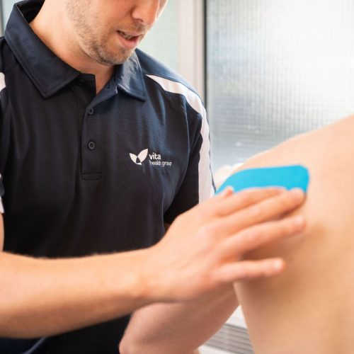 A healthcare professional applies a blue therapeutic tape to a patient's shoulder in a clinic. The professional wears a Vita Health Group polo shirt, beside a frosted glass window.