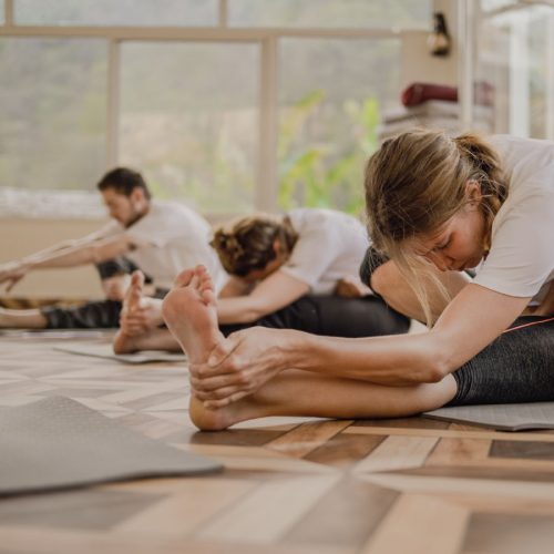 A person stretches on a yoga mat in a bright, spacious studio, with another person in a similar pose nearby. Light streams through large windows, creating a calm atmosphere.