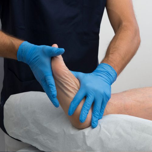 A healthcare professional wearing blue gloves examines a patient's foot, gently manipulating it while the patient lies on a treatment table covered in a white sheet.