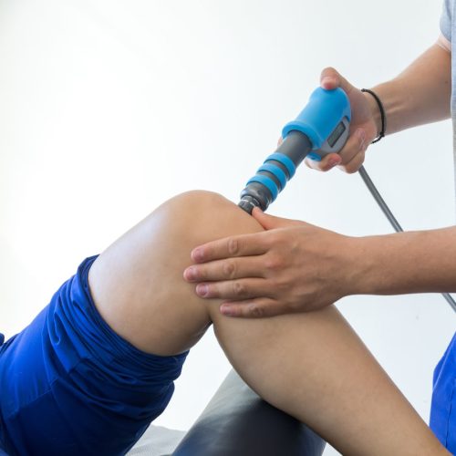A hand holds a blue therapy device, applying treatment to a person's knee in a clinical setting. The patient is wearing blue shorts, lying back on an examination table.