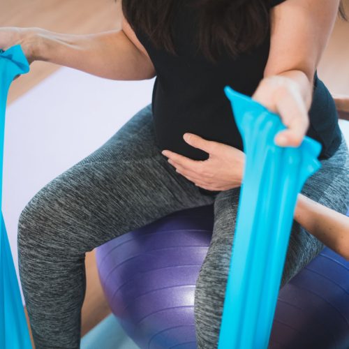 A person exercises on a purple stability ball, using a blue resistance band. They are in a fitness studio setting, wearing a black shirt and gray pants.