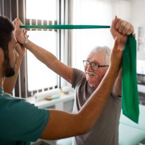 An elderly man exercises with a green resistance band, assisted by a healthcare professional, in a bright room with large windows.