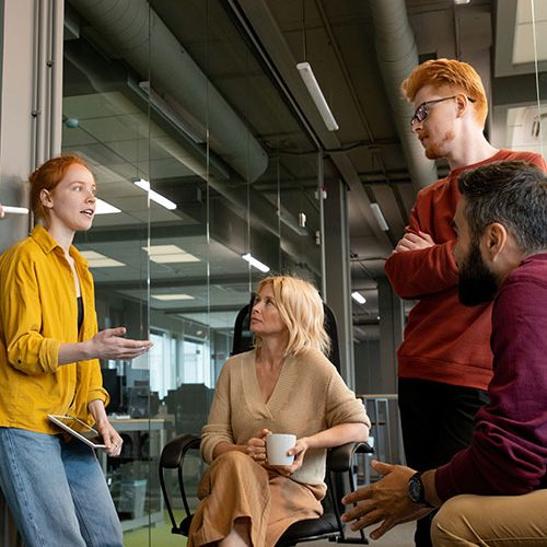 A group of four colleagues engage in discussion inside a modern office, with glass walls and overhead lighting. One person stands holding a notebook, while the others are seated, listening attentively.