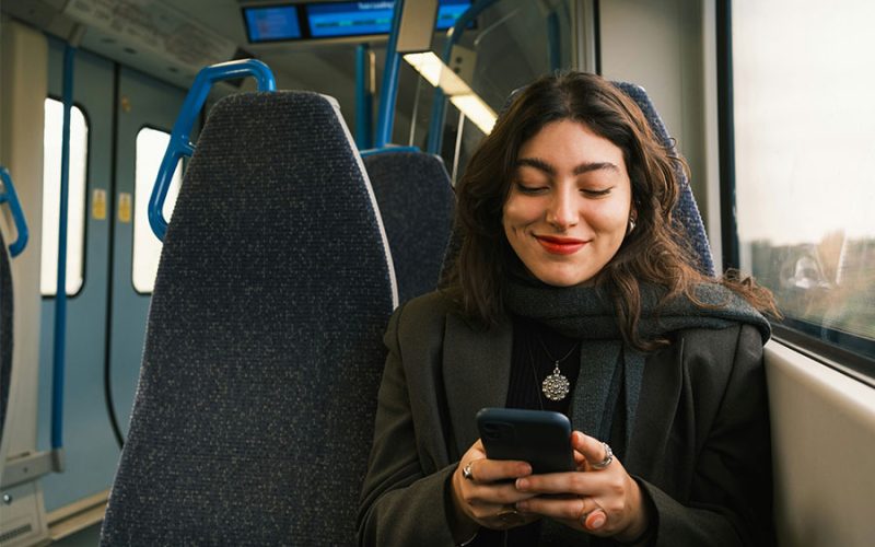 A person smiles while looking at a smartphone on a train. They are seated next to a window, wearing a scarf and jacket, with empty seats around them.