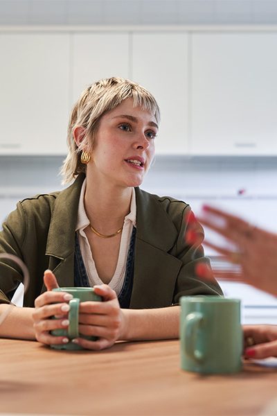 A person holding a green mug is sitting at a table, engaged in conversation with someone gesturing, in a kitchen setting with white cabinets.