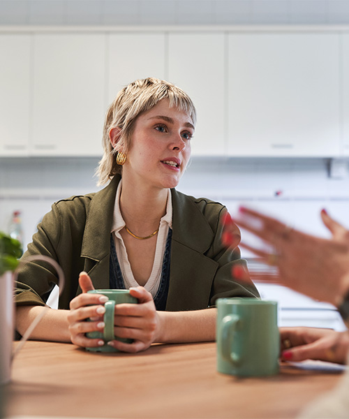 A person holding a green mug is sitting at a table, engaged in conversation with someone gesturing, in a kitchen setting with white cabinets.