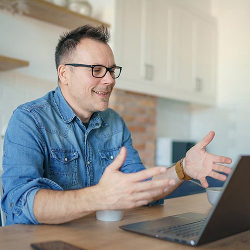 A man wearing glasses and a denim shirt gestures while talking to a laptop screen, sitting at a wooden table in a modern kitchen environment.