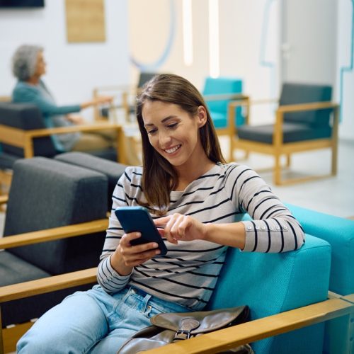 A woman is sitting on a modern chair using a smartphone, smiling. In the background, an older person is seated, creating a relaxed waiting room atmosphere.