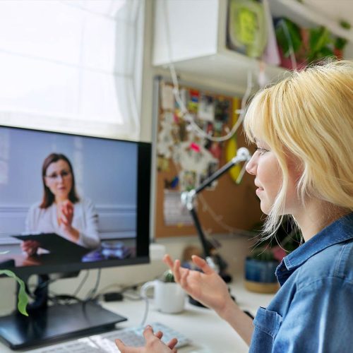 A person engages in a video call on a computer monitor, gesturing with hands, in a cluttered home office with a pinboard and plants in the background.