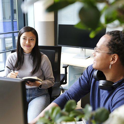 A woman holds a notebook, smiling and conversing with a man wearing headphones. They are seated in a modern office, surrounded by computer monitors and greenery.