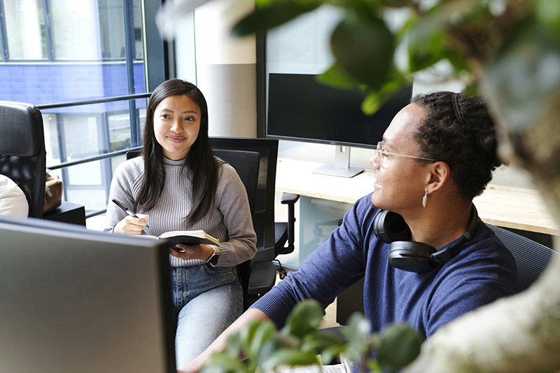 A woman holds a notebook, smiling and conversing with a man wearing headphones. They are seated in a modern office, surrounded by computer monitors and greenery.