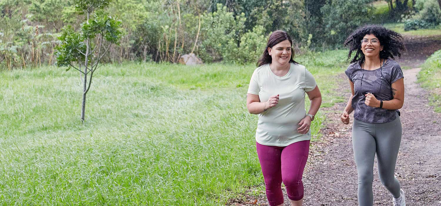 Two women are jogging along a park trail, surrounded by green trees and grass. One wears a white shirt and pink pants, while the other wears a patterned top and gray leggings.