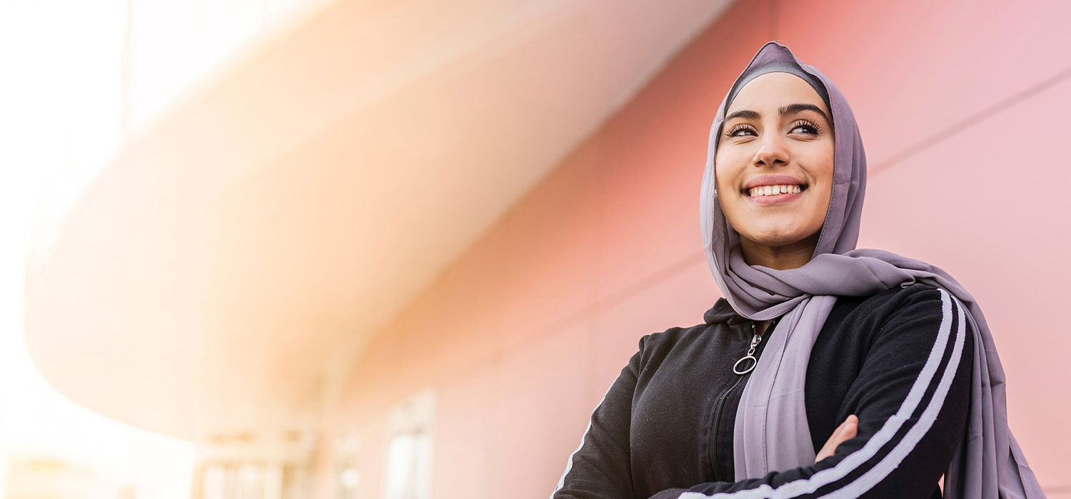 A person in a hijab smiles confidently, standing with crossed arms. The background features a modern, curved architectural structure with a soft pink color gradient. No text is present.