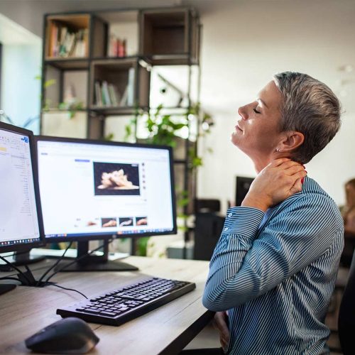 A person, sitting at a desk, massages their neck with a pained expression. They face two computer monitors displaying images and documents. Bookshelves and plants are in the background.