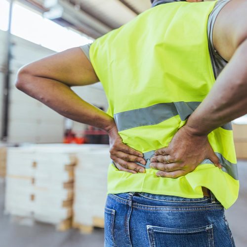 A person in a high-visibility vest holds their lower back in apparent discomfort, standing in a warehouse environment with stacked boxes in the background.