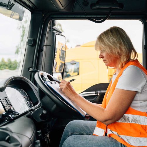 A person in a high-visibility vest is writing on a clipboard inside a truck cab, with another yellow vehicle visible outside the window.