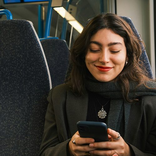 A person is sitting on a train, smiling at a smartphone. They are wearing a dark scarf and coat, with blue seats visible in the background.