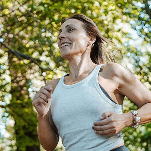A person is jogging outdoors, smiling with determination, wearing a white tank top. Green, sunlit trees form the background, suggesting a warm, pleasant day.