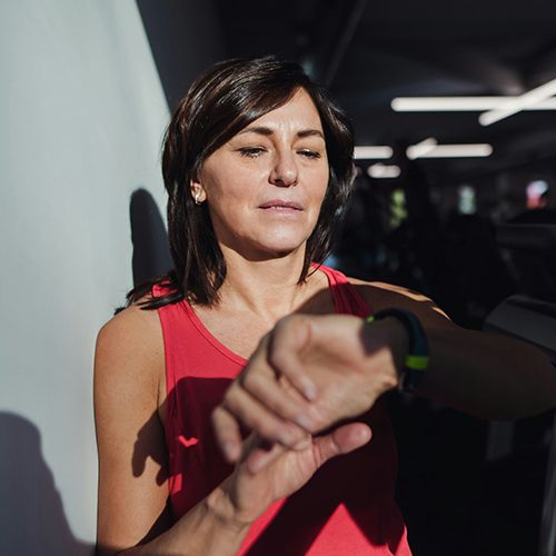 A person in a red tank top checks their smartwatch, standing in a well-lit gym with exercise equipment and fluorescent lights visible in the background.