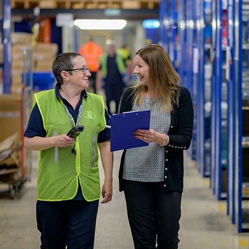 Two colleagues walk through a warehouse aisle, one holding a phone, the other a clipboard. They appear to be in conversation, surrounded by shelves filled with various items.