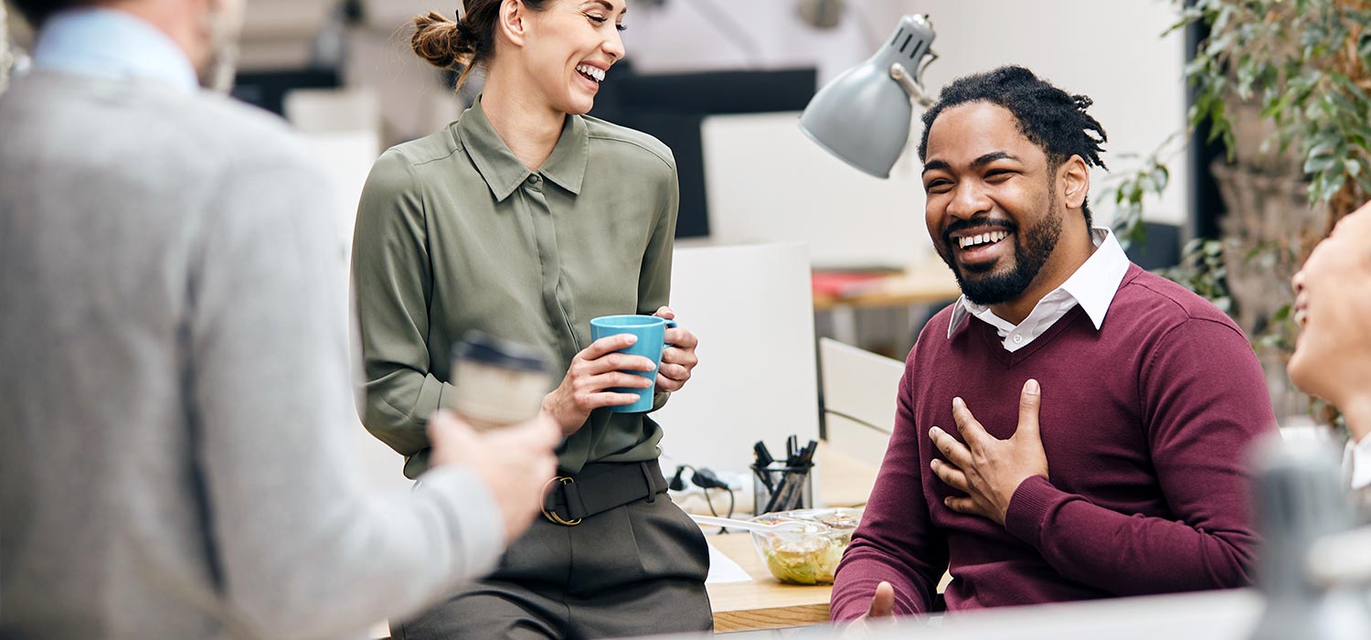 A group of colleagues is sitting and standing around a table, smiling and holding coffee cups, in a well-lit office space with plants in the background.