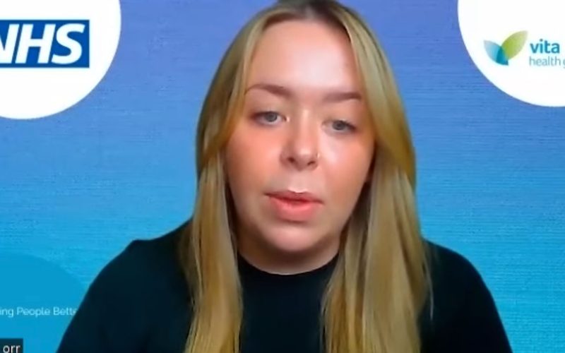 A screen capture of webinar where a woman with long blonde hair is speaking, facing forward against a blue background featuring NHS and Vita Health Group logos. Small, barely visible text reads, "Making People Better."