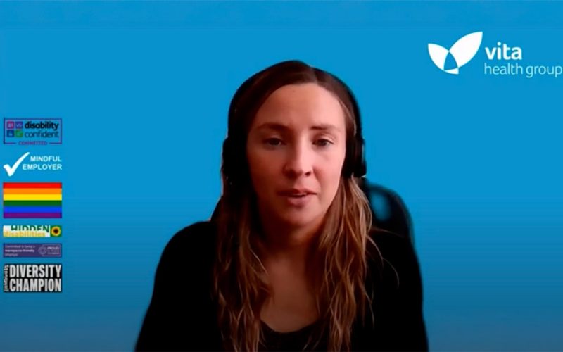 A screen capture of webinar where a woman wearing headphones speaks against a blue background. The Vita Health Group logo is in the top right corner. On the left are logos: "Disability Confident Committed," "Mindful Employer," a rainbow flag, "Hidden," and "Diversity Champion."